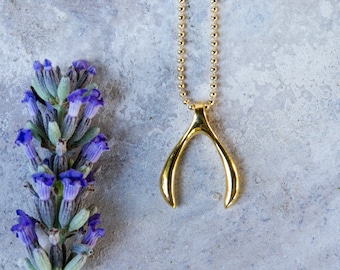 May include: A gold wishbone necklace on a chain with a grey background and a sprig of lavender.