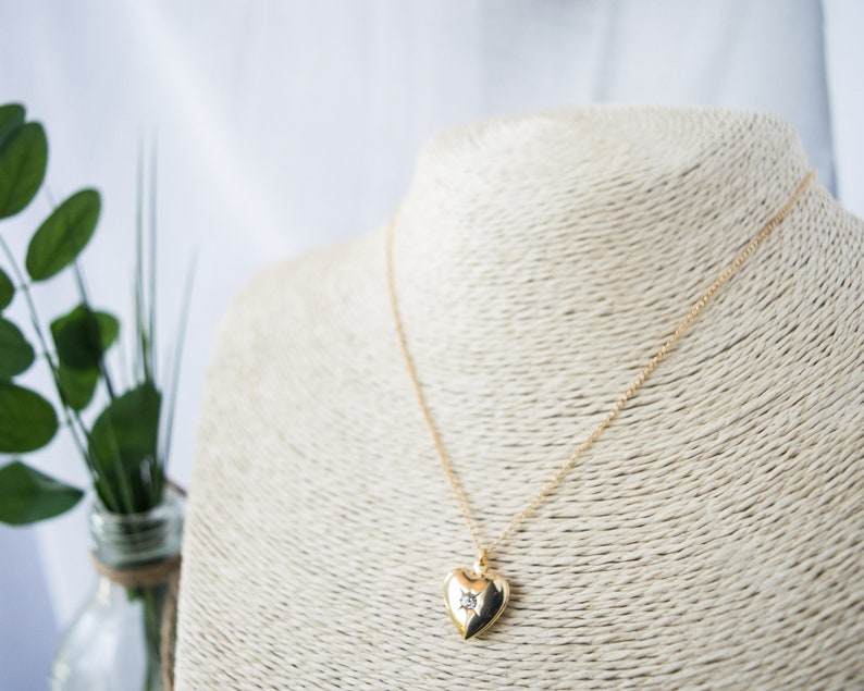 Pode incluir: Um colar de ouro com um pingente em forma de cora&ccedil;&atilde;o com um pequeno diamante no centro. O colar est&aacute; em um expositor branco.