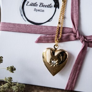Pode incluir: Colar com pingente de cora&ccedil;&atilde;o de ouro com um pequeno diamante no centro. O colar est&aacute; sobre um fundo branco com uma fita rosa.