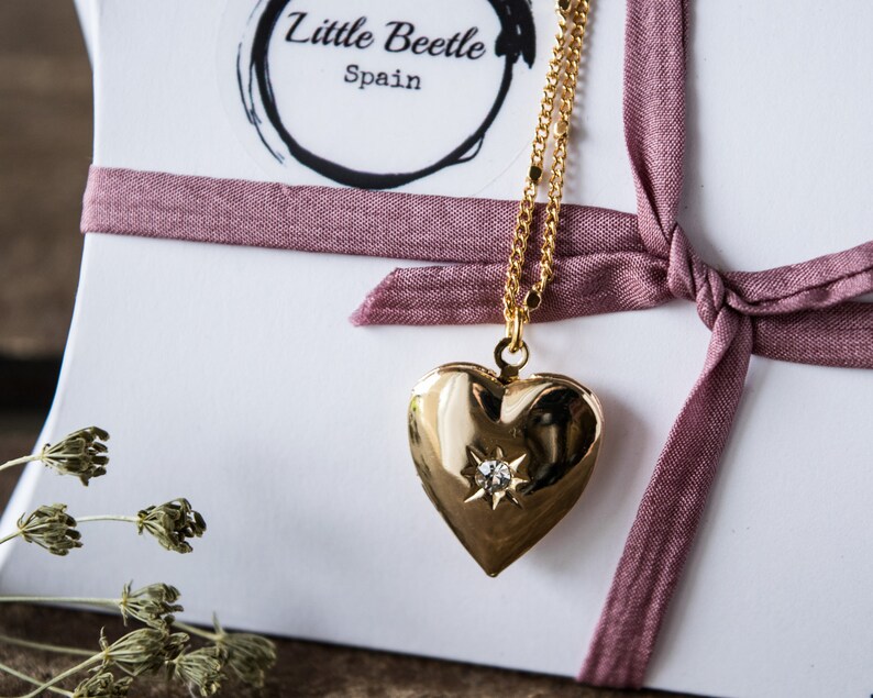 Pode incluir: Colar com pingente de cora&ccedil;&atilde;o de ouro com um pequeno diamante no centro. O colar est&aacute; sobre um fundo branco com uma fita rosa.