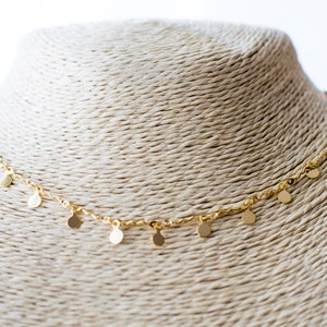 Peut inclure: Un collier en chaîne dorée avec de petits pendentifs ronds. Le collier est présenté sur un mannequin beige texturé.
