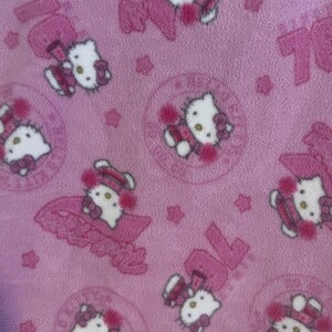 May include: Pink fabric featuring a repeating pattern of the Hello Kitty character in various poses. The design includes the number 70 and star shapes, all in shades of pink and white. The fabric has a soft, plush texture.