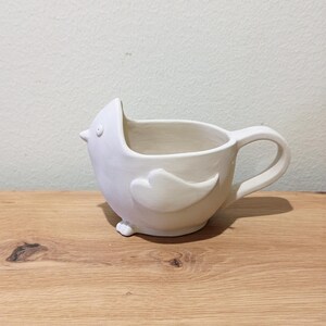May include: A matte white ceramic mug shaped like a bird. The mug features a pointed beak, a small circular eye, a raised wing detail on the side, and a curved handle. The bird-shaped vessel has an open top and a smooth, unglazed appearance.