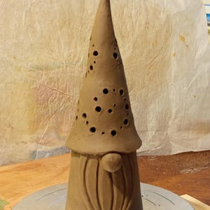 K&ouml;nnte beinhalten: Eine hohe, kegelf&ouml;rmige Tonskulptur einer Wichtelfigur mit einer spitzen M&uuml;tze, die zahlreiche kreisf&ouml;rmige Ausschnitte aufweist. Die Figur hat eine glatte, unglasierte braune Oberfl&auml;che, eine runde Nase und einen langen, strukturierten Bart.