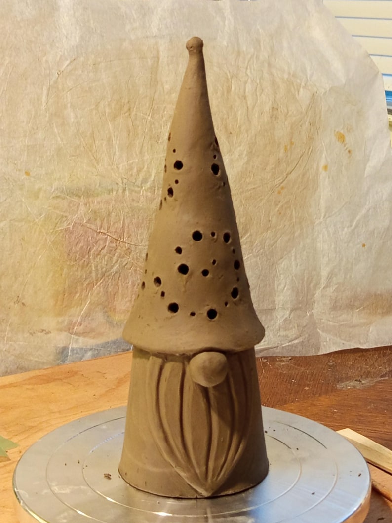 K&ouml;nnte beinhalten: Eine hohe, kegelf&ouml;rmige Tonskulptur einer Wichtelfigur mit einer spitzen M&uuml;tze, die zahlreiche kreisf&ouml;rmige Ausschnitte aufweist. Die Figur hat eine glatte, unglasierte braune Oberfl&auml;che, eine runde Nase und einen langen, strukturierten Bart.