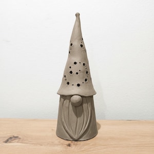 K&ouml;nnte beinhalten: Eine hohe, kegelf&ouml;rmige Keramik-Gnomfigur in mattem Grau. Der Gnom hat eine spitze M&uuml;tze mit zahlreichen kreisf&ouml;rmigen Aussparungen unterschiedlicher Gr&ouml;&szlig;e, eine runde Nase und einen modellierten Bart mit vertikalen Linien am zylindrischen Sockel.