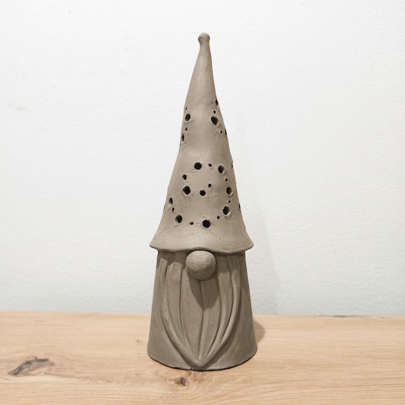 K&ouml;nnte beinhalten: Eine hohe, kegelf&ouml;rmige Keramik-Gnomfigur in mattem Grau. Der Gnom hat eine spitze M&uuml;tze mit zahlreichen kreisf&ouml;rmigen Aussparungen unterschiedlicher Gr&ouml;&szlig;e, eine runde Nase und einen modellierten Bart mit vertikalen Linien am zylindrischen Sockel.