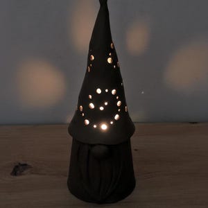 K&ouml;nnte beinhalten: Eine matte schwarze Keramik-Gnomfigur mit einer hohen, spitzen M&uuml;tze, die kreisf&ouml;rmige Ausschnitte aufweist. Licht scheint durch die L&ouml;cher in der M&uuml;tze und erzeugt einen leuchtenden Effekt. Der Gnom hat eine runde Nase und einen strukturierten Bart.