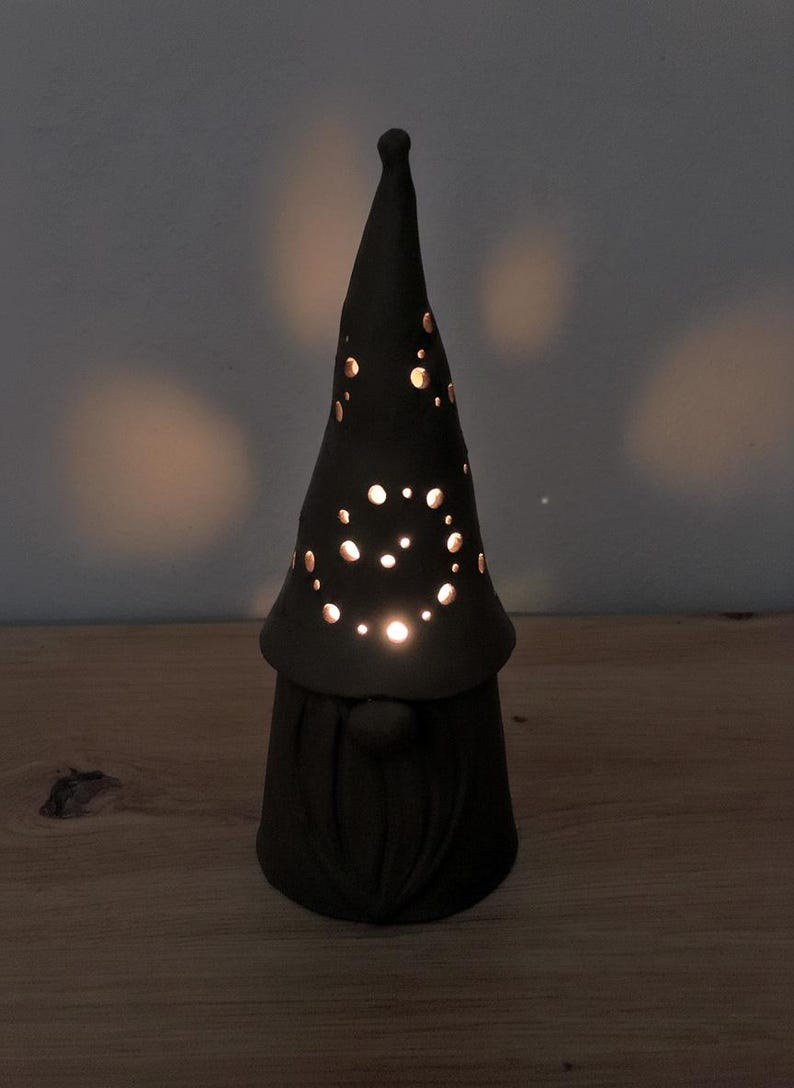 K&ouml;nnte beinhalten: Eine matte schwarze Keramik-Gnomfigur mit einer hohen, spitzen M&uuml;tze, die kreisf&ouml;rmige Ausschnitte aufweist. Licht scheint durch die L&ouml;cher in der M&uuml;tze und erzeugt einen leuchtenden Effekt. Der Gnom hat eine runde Nase und einen strukturierten Bart.