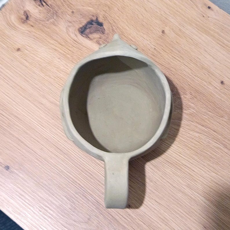 May include: A light beige ceramic mug with a rounded, bowl-like body and a single handle. The mug features a pointed, snout-like protrusion at the top rim, giving it a distinct animal-inspired shape. The interior is smooth and unglazed.