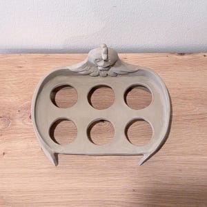 Op de afbeelding: Een lichtgrijze keramische eierhouder met een gebeeldhouwde kippenfiguur aan de bovenzijde. Het rechthoekige dienblad heeft zes ronde uitsparingen in twee rijen van drie, ontworpen om individuele eieren stevig in een rechtopstaande positie te houden.