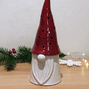 K&ouml;nnte beinhalten: Eine Keramik-Gnom-Figur mit einer hohen, gl&auml;nzend roten M&uuml;tze, die mit verstreuten kreisf&ouml;rmigen Aussparungen versehen ist, und einer glatten wei&szlig;en Basis. Der Gnom hat einen markanten wei&szlig;en Bart und eine runde Nase, ein dekoratives Objekt.