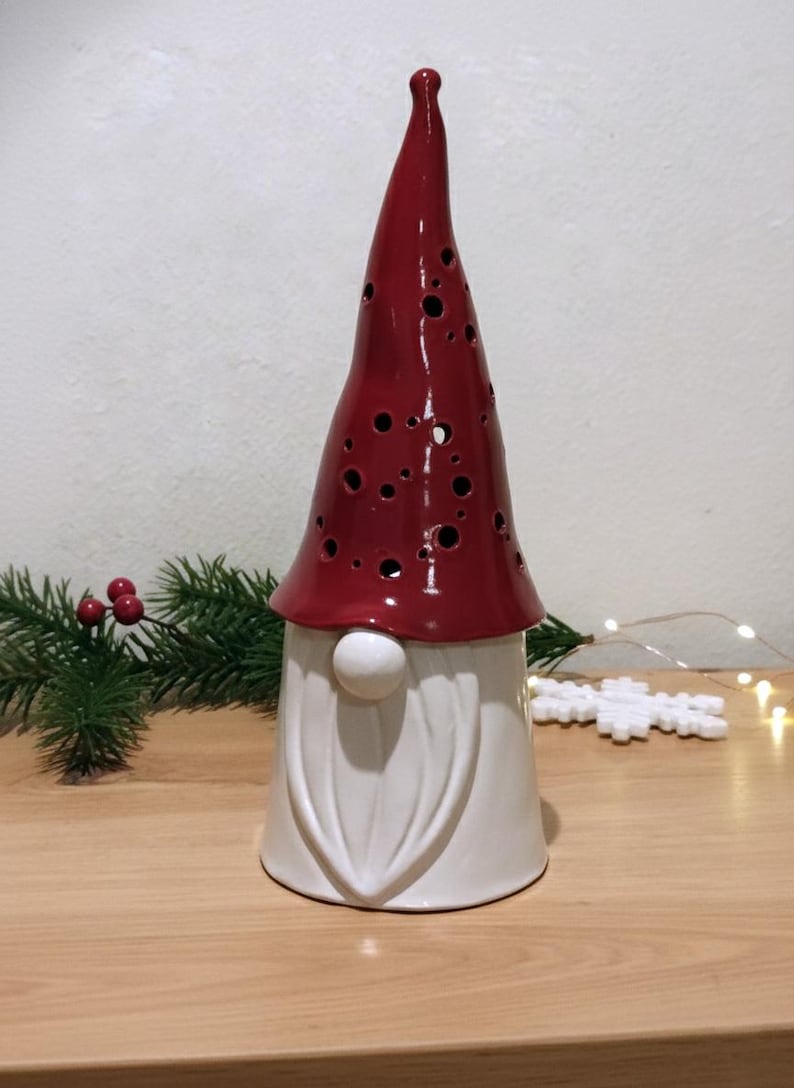 K&ouml;nnte beinhalten: Eine Keramik-Gnom-Figur mit einer hohen, gl&auml;nzend roten M&uuml;tze, die mit verstreuten kreisf&ouml;rmigen Aussparungen versehen ist, und einer glatten wei&szlig;en Basis. Der Gnom hat einen markanten wei&szlig;en Bart und eine runde Nase, ein dekoratives Objekt.