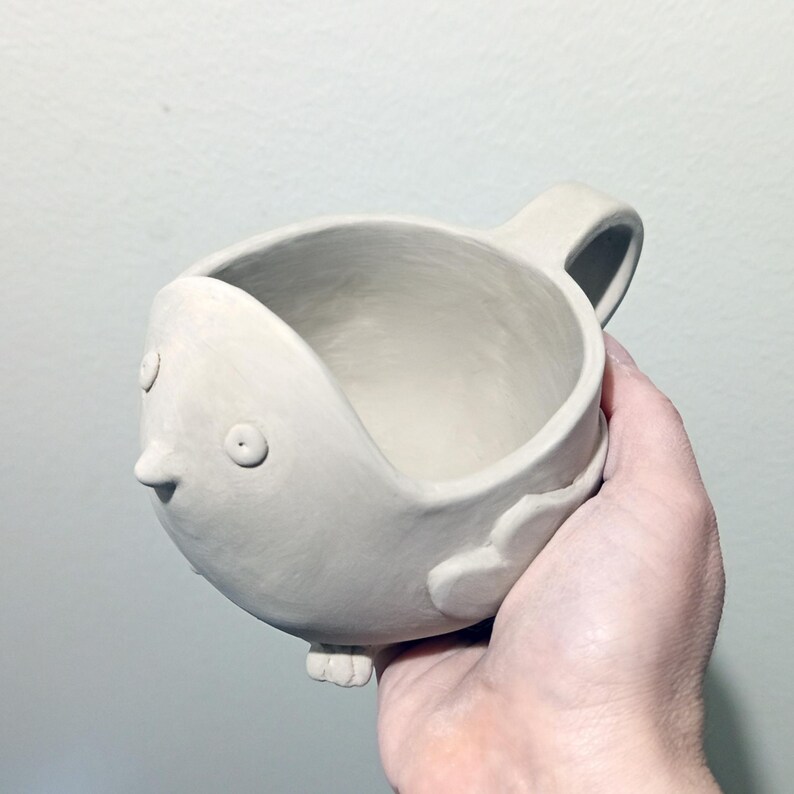 May include: A matte off-white ceramic mug shaped like a stylized bird. The mug features a rounded body, a small pointed beak, two circular eyes, a handle on the side, and small sculpted feet at the base. The surface has a smooth, hand-formed texture.