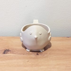 May include: A matte off-white ceramic mug shaped like a bird with a pointed beak, two small circular eyes, and side protrusions resembling wings. The vessel features a small handle at the top and stands on two small feet, with a smooth finish.