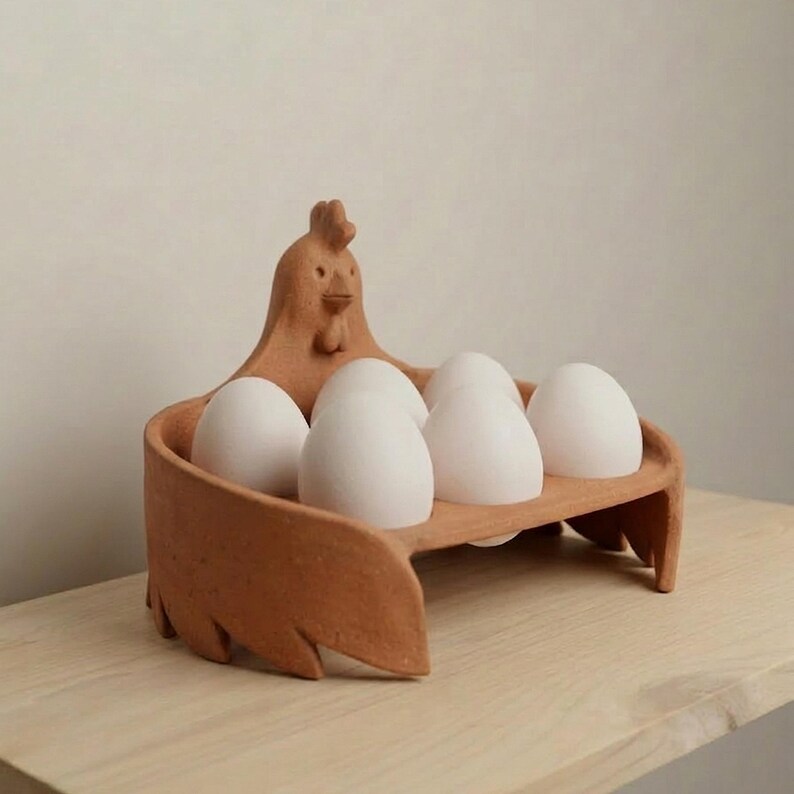 Op de afbeelding: Een terracotta-kleurige keramische eierhouder in de vorm van een kip met een getextureerd oppervlak. De houder heeft een centrale figuur van een kip met een kam en snavel en bevat zes gladde, witte, ovale eieren in twee rijen.