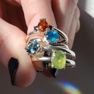 May include: Four silver rings with colorful gemstones. The rings are stacked on a finger. The gemstones are orange, blue, blue, and green.