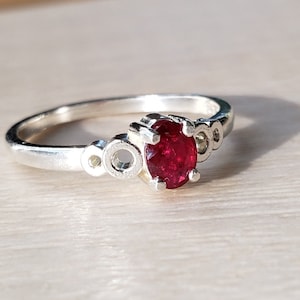 May include: A silver ring with a red oval gemstone set in a prong setting. The band features a decorative design of small circles.