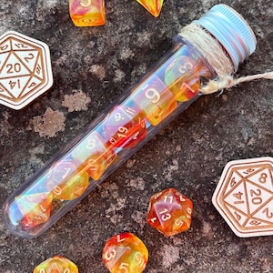 May include: A clear plastic tube filled with orange and yellow iridescent dice. The tube is tied with twine and has a white cap. There are several dice scattered around the tube, including a wooden 20-sided die with the numbers 8, 20, 14, and 2 printed on it.