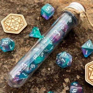 May include: A clear glass test tube filled with iridescent blue and green dice. The test tube is tied with twine and has a cork stopper. The dice are scattered around the test tube on a brown, rocky surface.