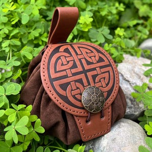 May include: A brown suede pouch with a leather flap and handle. The flap features a Celtic knot design and a bronze-colored button. The pouch is set against a backdrop of green foliage and rocks.