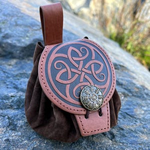 May include: A brown leather pouch with a Celtic knot design on the flap. The pouch has a suede-like texture and a small handle. A metal button secures the flap. The pouch is likely used for carrying small items.