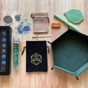 May include: An assortment of tabletop gaming accessories, including dice, a dice tray, and small pouches. The image features a black dice case, a tube of dice, a black bag with a yellow 20 symbol, and a green dice tray.