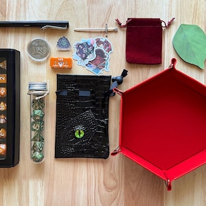May include: A collection of gaming accessories, including dice sets, a dice tray, a pen, stickers, a coin, and a small velvet bag. The dice sets are in a black case and a glass tube. The dice tray is red.