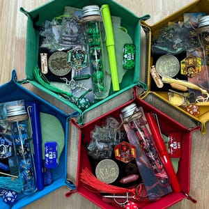May include: A collection of colorful hexagonal dice trays filled with various gaming accessories. Each tray, in green, yellow, red, and blue, contains dice, pens, coins, and other game-related items. The trays are arranged on a light wood surface.
