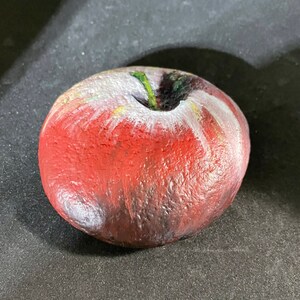 May include: A hand-painted, red apple-shaped decorative stone. The apple features white and yellow highlights, a green stem, and a dark shaded area at the top. The stone is set against a dark background.