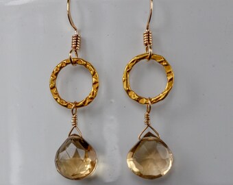 May include: A pair of gold-colored earrings with a hammered texture. Each earring features a circular design and a teardrop-shaped, light brown gemstone. The earrings have a hook closure.