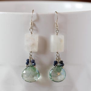 May include: A pair of dangle earrings with silver hooks. Each earring features a white, square-shaped stone, a cluster of small blue beads, and a large, faceted, teardrop-shaped green stone. The earrings are displayed against a white background.