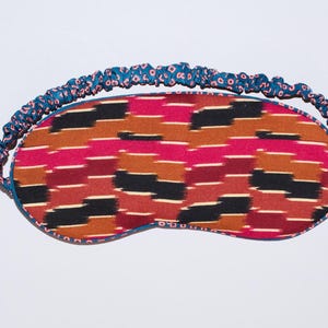 May include: A patterned sleep mask with a blue elastic band. The mask features a design of horizontal stripes in shades of red, brown, and black. The elastic band is blue with a repeating pink and blue pattern.
