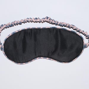May include: A black sleep mask with a patterned pink and blue elastic band. The mask is oval-shaped and made of a dark, possibly silk-like material. The band is gathered and features a repeating floral design.