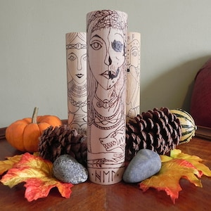 May include: Three wooden cylinders with black and white illustrations of Norse deities. The cylinder in the center features a woman with a skull face and the text "NMM". The cylinder on the left features a woman with a crown and the text "MMS". The cylinder on the right features a man with a beard and a helmet.