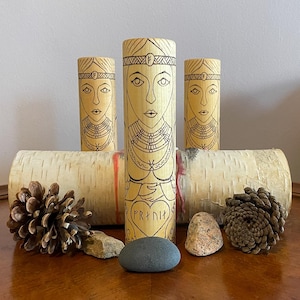 May include: Three wooden cylinders with a design of a woman's face and a heart with the word "FRYNJA" inscribed on the front. The cylinders are sitting on a birch bark surface with pine cones and stones.