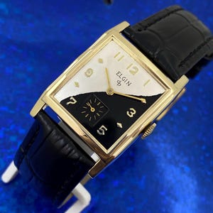 Peut inclure: Une montre Elgin vintage avec un bo&icirc;tier rectangulaire dor&eacute; et un bracelet en cuir noir. Le cadran de la montre est divis&eacute; en diagonale, avec un design blanc et noir. La montre a des aiguilles et des index dor&eacute;s, avec la marque "ELGIN".