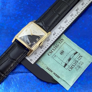Peut inclure: Une montre Elgin vintage avec un bo&icirc;tier rectangulaire dor&eacute; et un bracelet en cuir noir. Le cadran est divis&eacute; en sections noires et blanches avec des aiguilles et des chiffres dor&eacute;s. Une r&egrave;gle et un paquet vert sont &eacute;galement visibles.