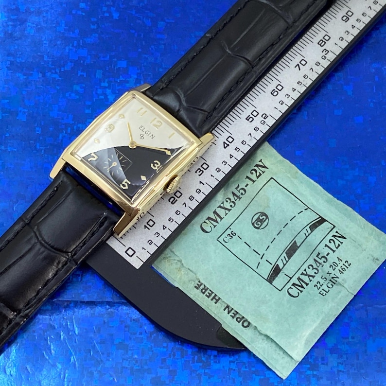 Peut inclure: Une montre Elgin vintage avec un bo&icirc;tier rectangulaire dor&eacute; et un bracelet en cuir noir. Le cadran est divis&eacute; en sections noires et blanches avec des aiguilles et des chiffres dor&eacute;s. Une r&egrave;gle et un paquet vert sont &eacute;galement visibles.