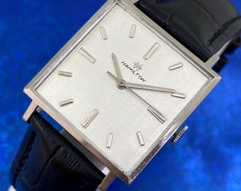 May include: A vintage Hamilton wristwatch with a square silver face and a black leather band. The watch face has silver hour markers and hands. The word "HAMILTON" is printed on the watch face.