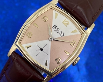 Könnte beinhalten: Vintage Bulova-Uhr mit goldfarbenem Gehäuse und braunem Lederarmband. Das Zifferblatt ist in vier Abschnitte aus Weiß und Rosa unterteilt, mit goldfarbenen Ziffern und Zeigern. Der Text "Bulova 17 Jewels" ist sichtbar.