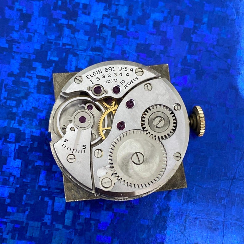 Peut inclure: Gros plan d'un mouvement de montre Elgin, avec des engrenages, des bijoux et le texte "ELGIN 681 U-S-A". Le m&eacute;canisme est argent&eacute; avec des engrenages dor&eacute;s et une couronne textur&eacute;e. Fond bleu textur&eacute;.