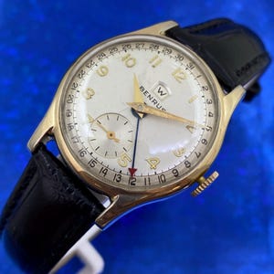 Może przedstawiać: Złoty zegarek na rękę Benrus z kremową tarczą i czarnym skórzanym paskiem. Zegarek posiada złote wskazówki i indeksy godzinowe, małą tarczę sekundową i okienko daty. Na tarczy widnieje nazwa marki i szereg cyfr.