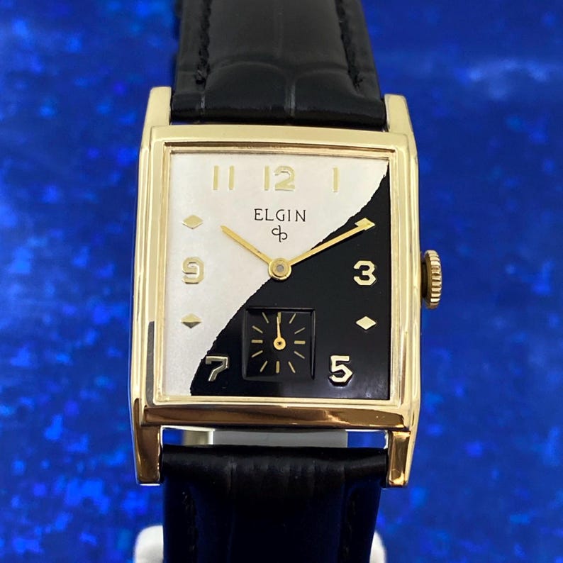 Peut inclure: Une montre Elgin vintage avec un bo&icirc;tier rectangulaire dor&eacute; et un bracelet en cuir noir. Le cadran est divis&eacute; en diagonale, avec un c&ocirc;t&eacute; blanc portant les chiffres 11, 12 et 1, et un c&ocirc;t&eacute; noir avec les chiffres 3 et 5. La montre a un petit cadran de secondes.