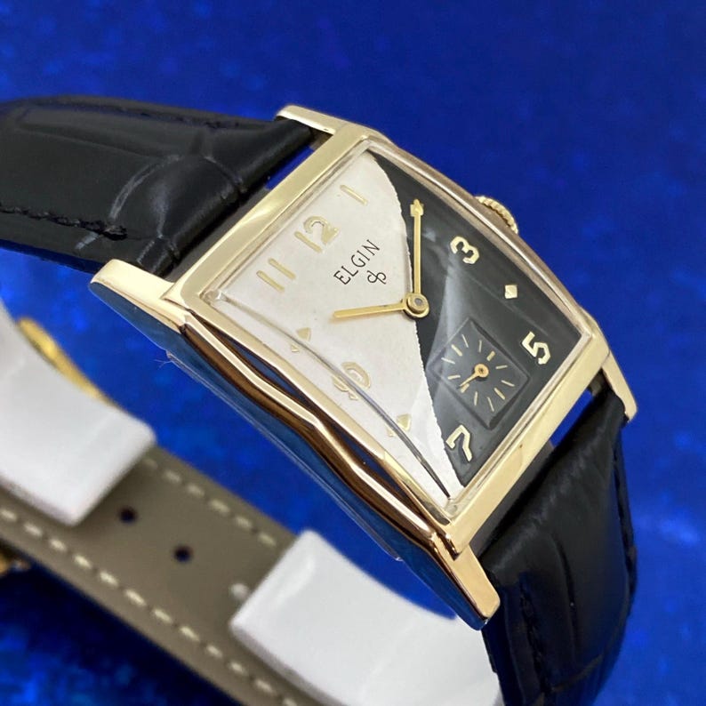 Peut inclure: Une montre Elgin vintage avec un bo&icirc;tier rectangulaire dor&eacute; et un bracelet en cuir noir. Le cadran est divis&eacute;, avec un c&ocirc;t&eacute; blanc et un c&ocirc;t&eacute; noir. Le c&ocirc;t&eacute; blanc affiche le logo Elgin et des aiguilles dor&eacute;es. Le c&ocirc;t&eacute; noir a des chiffres dor&eacute;s.
