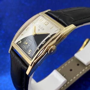 Peut inclure: Une montre Elgin vintage avec un bo&icirc;tier dor&eacute; et un bracelet en cuir noir. Le cadran est divis&eacute; en sections noires et blanches, avec des aiguilles et des chiffres dor&eacute;s. La montre a une forme rectangulaire et un design classique.