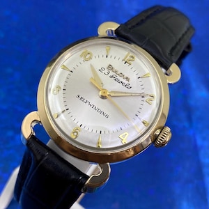 Puede incluir: Un reloj de pulsera automático Bulova vintage con caja dorada y correa de cuero negro. La esfera del reloj es blanca con agujas y números dorados. El reloj indica "Bulova 23 Jewels".