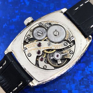 Pu&ograve; includere: Il retro di un orologio Gruen Guild argentato con un cinturino in pelle nera. Il quadrante dell'orologio &egrave; aperto, rivelando il meccanismo interno dell'orologio. L'orologio ha un movimento svizzero ed &egrave; contrassegnato da "Fifteen Jewels" e "Three Adj."