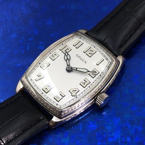 Pu&ograve; includere: Un orologio da polso Gruen in argento con quadrante bianco e lancette nere. L'orologio ha una cassa rettangolare e un cinturino in pelle nera.