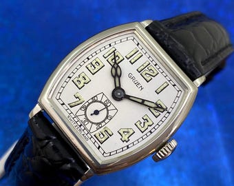 May include: A vintage Gruen wristwatch with a rectangular silver case and a white face. The watch features black hands, black Arabic numerals, and a small seconds dial. The black leather band complements the watch's classic design. The watch is marked "SWITZERLAND".